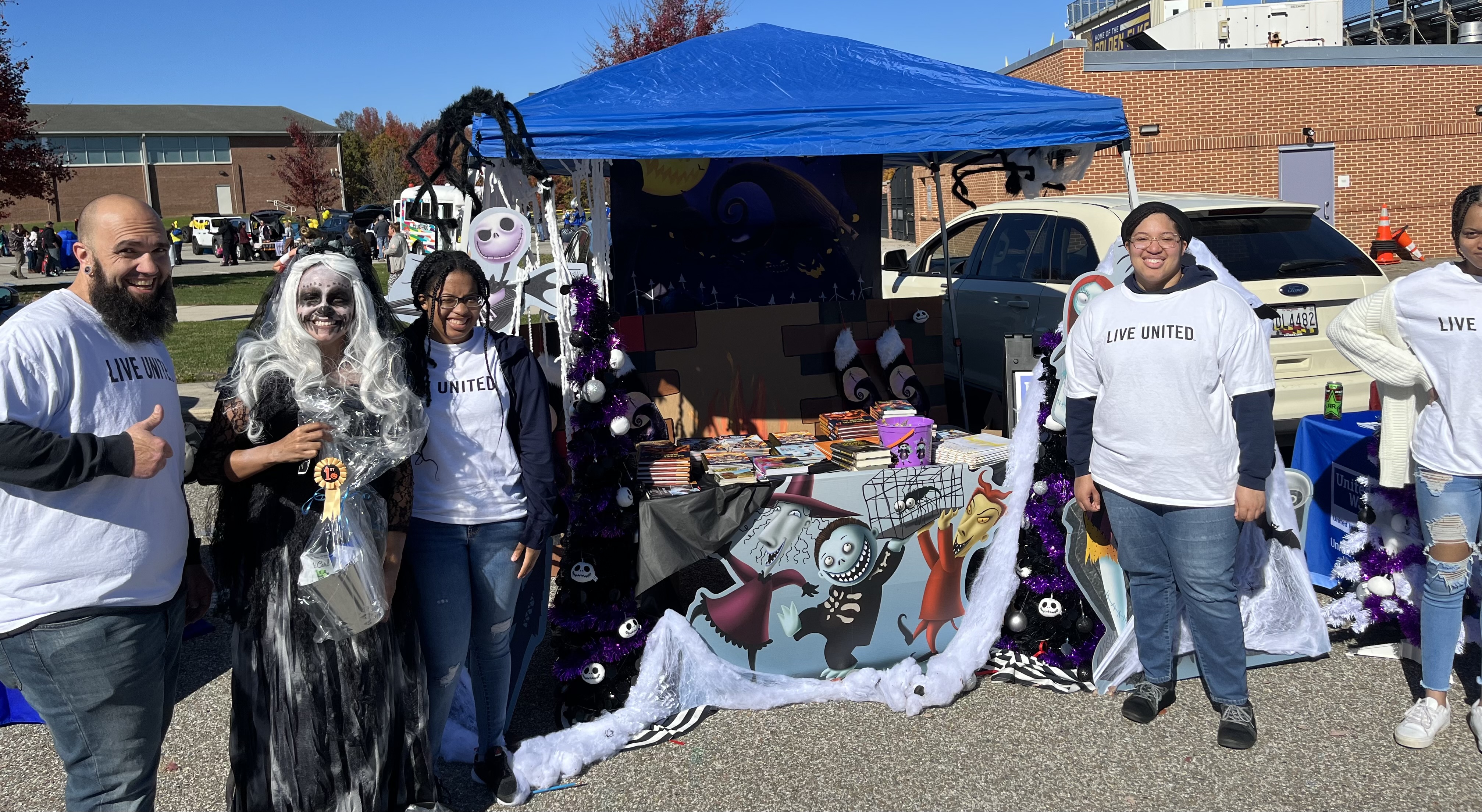 5 people with no hair, brown hair and brown eyes wearing white shirts with black lettering that says Live United. Blue tent in background with school and white car.